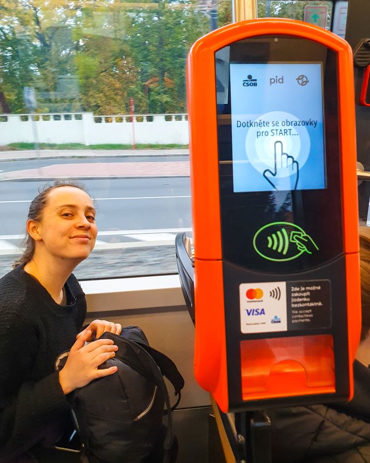 Prague Czechia A Complete Guide to Prague Public Transport Krissie smiling while sitting down on a tram in Prague, Czechia. On her left is a red and black ticket machine with spots to tap your payment card and then for your ticket to come out under it. A Complete Guide to Prague Public Transport