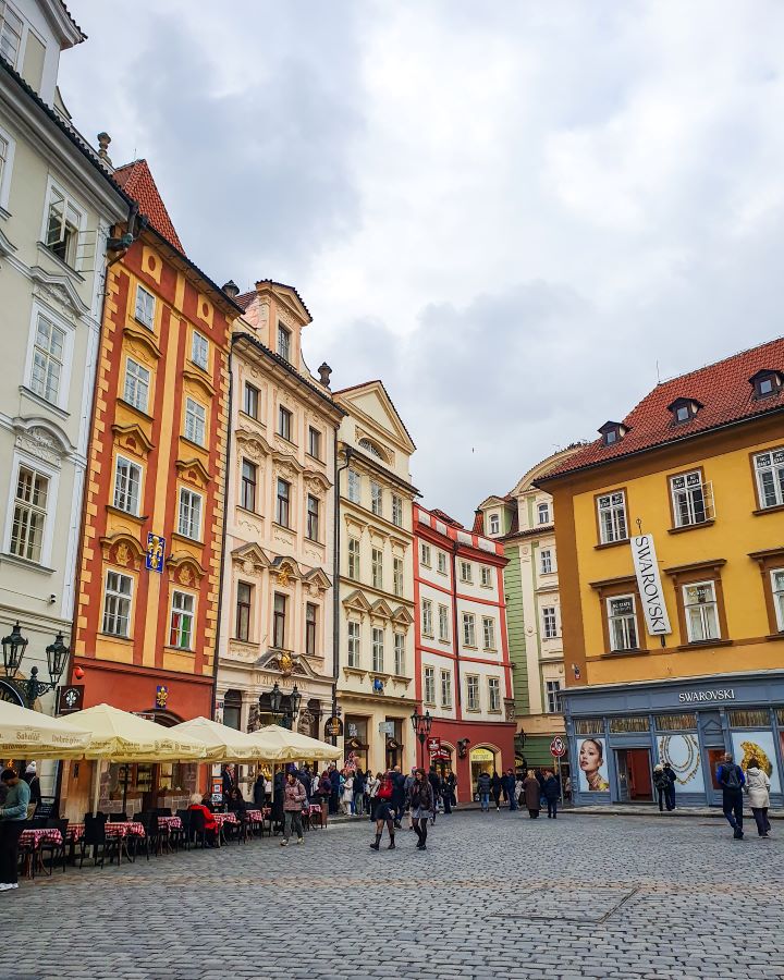 A town square in Prague, Czechia with buildings in yellow, orange, pink, red and green. They're multiple levels high with umbrellas out the front and people walking around. How To Visit Prague On a Budget: Best Three Day Itinerary