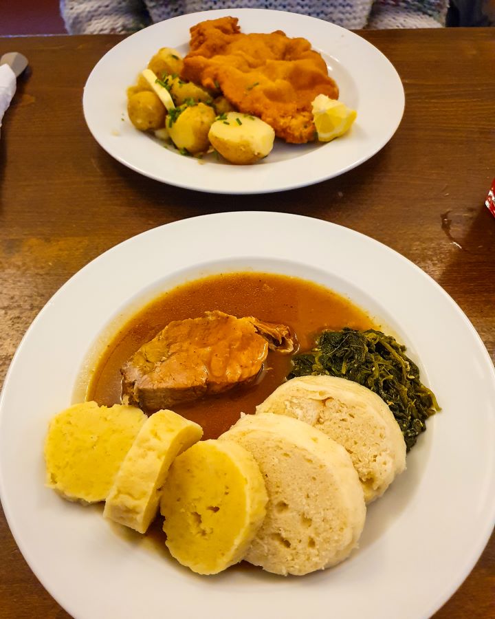 Two plates of food from Lokál Dlouhááá in Prague, Czechia. The front plate has round dumplings, a piece of pork, some spinach and some brown sauce on it and the back plate has a schnitzel and some potatoes. How To Visit Prague On a Budget: Best Three Day Itinerary