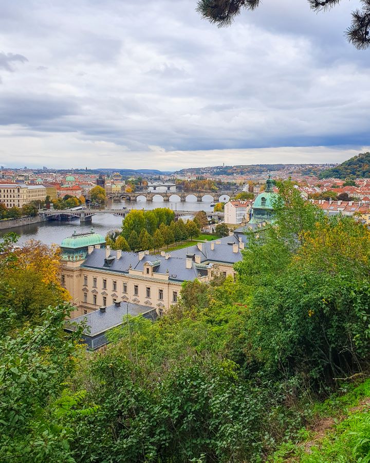 A view of Prague, Czechia from Letná Park in Prague, Czechia. There's green trees in the foreground and the river, bridges and buildings with orange roofs and green domes in the background. How To Visit Prague On a Budget: Best Three Day Itinerary