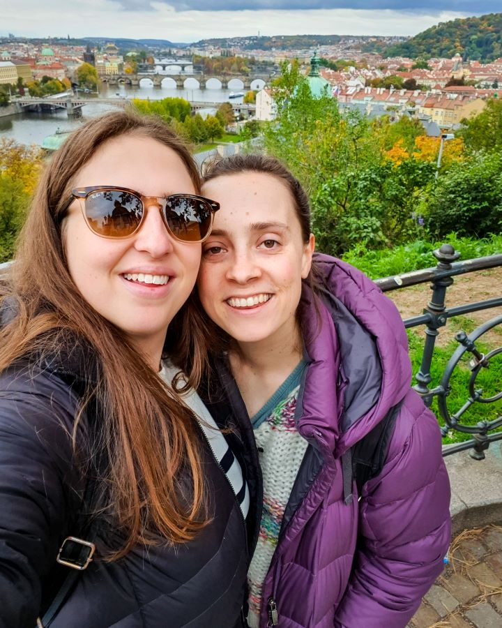 How To Visit Prague On a Budget: Best Three Day Itinerary. Katherine and Krissie smiling at the camera with a view of Prague, Czechia behind them. There's lots of green plants around with the Vltava river and lots of bridges going over it. There's also lots of buildings with orange roofs.