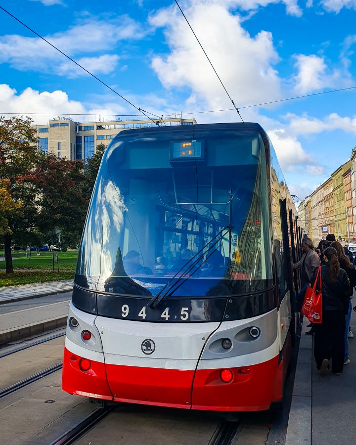 Prague Czechia A Complete Guide to Prague Public Transport A red, white and black tram in Prague, Czechia. There's tram lines above it, tracks under it and people on the right side waiting to get on. A Complete Guide to Prague Public Transport