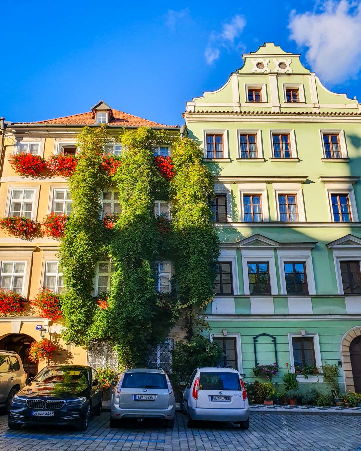 Two cute buildings in Lesser town, Prague, Czechia. The one on the left is covered in green vines and the one on the right is green with a curved top. There's cars parked out the front and the sky is blue. How To Visit Prague On a Budget: Best Three Day Itinerary
