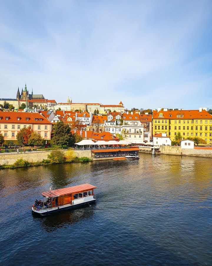 Charles Bridge Vltava River Prague Castle Czechia A Complete Guide to Prague Public Transport A little orange and white ferry on the river in Prague, Czechia. Behind the water are orange, white and yellow buildings with orange roofs. There's spots of trees and behind them is the brown castle on a hill above it all. A Complete Guide to Prague Public Transport