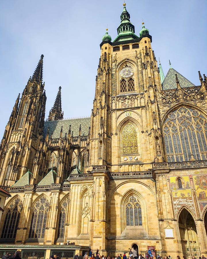 The incredible St. Vitus Cathedral in Prague, Czechia. The cathedral is sand coloured with lots of pointed bits, spires, a green roof and big stain glass windows. How To Visit Prague On a Budget: Best Three Day Itinerary