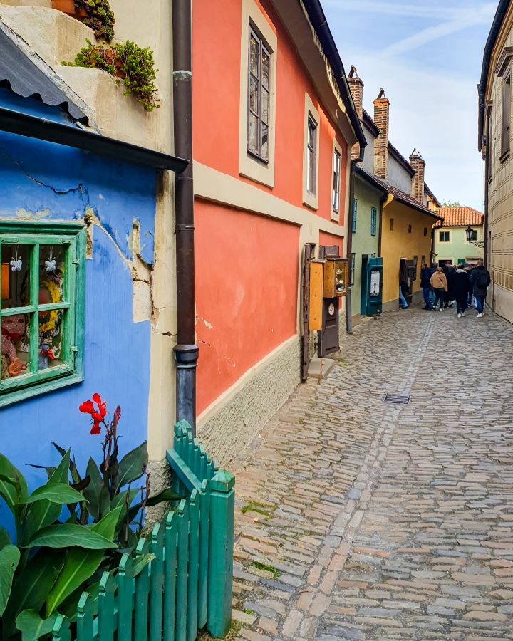 Golden Lane in Prague, Czechia. There's a cobbled street with blue, red, green and yellow small buildings on the left. The houses look older and out the front of one of them is a small green picket fence. How To Visit Prague On a Budget: Best Three Day Itinerary
