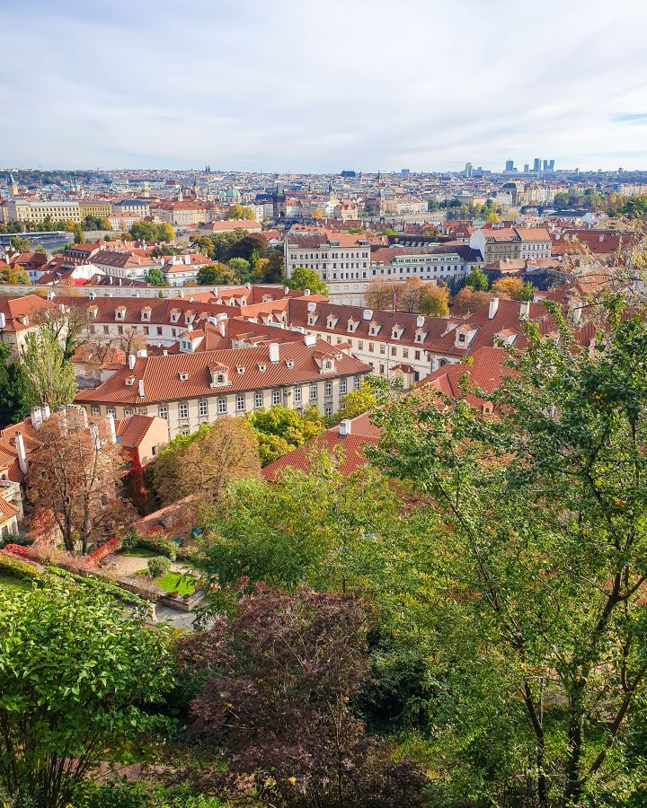 Prague Castle Czechia A Complete Guide to Prague Public Transport A Complete Guide to Prague Public Transport. In the foreground are green trees and behind them a view of Prague, Czechia. There's lots of white buildings with orange roofs with spots of green trees in between them all.