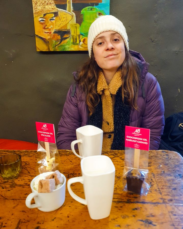 How to See Luxembourg City in a Day Chocolate House Krissie sitting across the table smiling at the camera in a café in Luxembourg City. On the table are two chocolate spoons, two tall mugs of milk and a cup with cream and marshmallows. How to See Luxembourg City in a Day