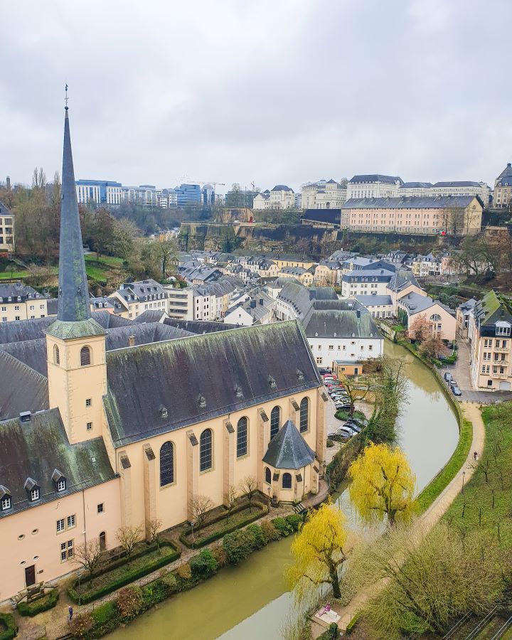 How to See Luxembourg City in a Day Christmas Market Ville Haute How to See Luxembourg City in a Day. A view over the Alzette river with a beige and grey church on the left and more grey and white and beige buildings in the background in the Gurnd in Luxembourg City. Around them are trees and the sky is grey and overcast.