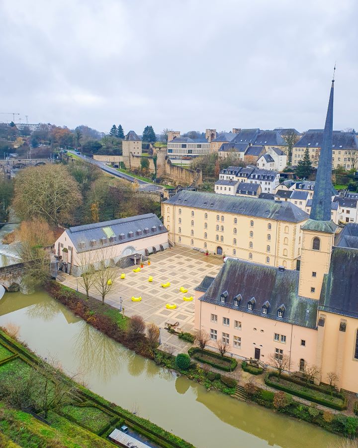 How to See Luxembourg City in a Day Christmas Market The Grund Looking down at Neumünster Abbey in the Grund in Luxembourg City. It's a beige building with a dark grey roof with one tall church spire. Behind it are other buildings and trees with a grey sky above it all. How to See Luxembourg City in a Day