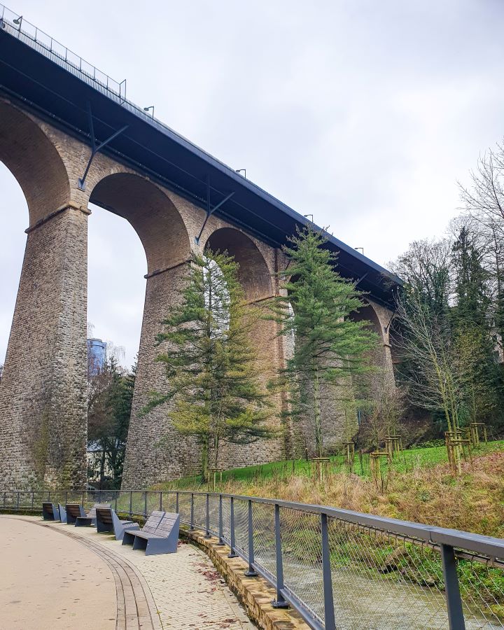 How to See Luxembourg City in a Day Christmas Market A path going under a stone viaduct bridge in Luxembourg City. On top of the bridge is a road and under it is green grass and trees. How to See Luxembourg City in a Day