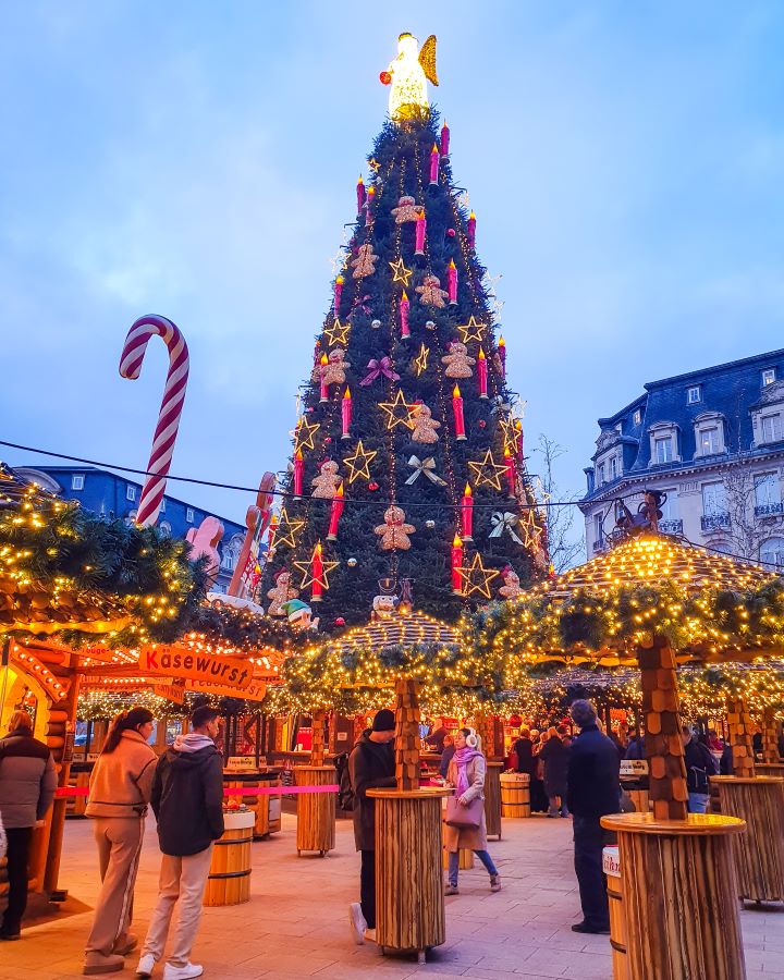 Little wooden stands with fairy lights on top with people walking around underneath at a market in Luxembourg City. Behind them is a large Christmas tree with candles, stars and gingerbread men decorations on it with a big angel on top. There's also a red and white candy cane on the left. Make the Most of Christmas in Luxembourg City