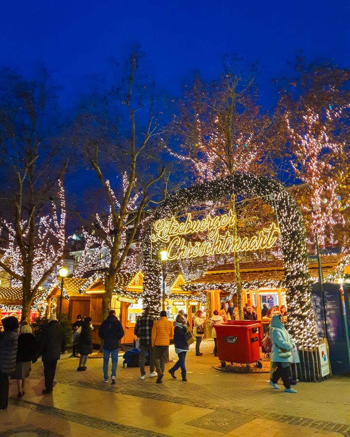 The entrance to the main Christmas market in Luxembourg City. There's lots of lights in the trees and wooden huts under them. There's also lots of people walking around and the sky is dark. Make the Most of Christmas in Luxembourg City