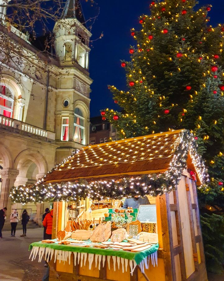 In a Christmas market in Luxembourg City. There's a hut with Christmas lights on it and it's selling crafts. Behind it is a Christmas tree with red and yellow lights and behind that is the town hall. Make the Most of Christmas in Luxembourg City
