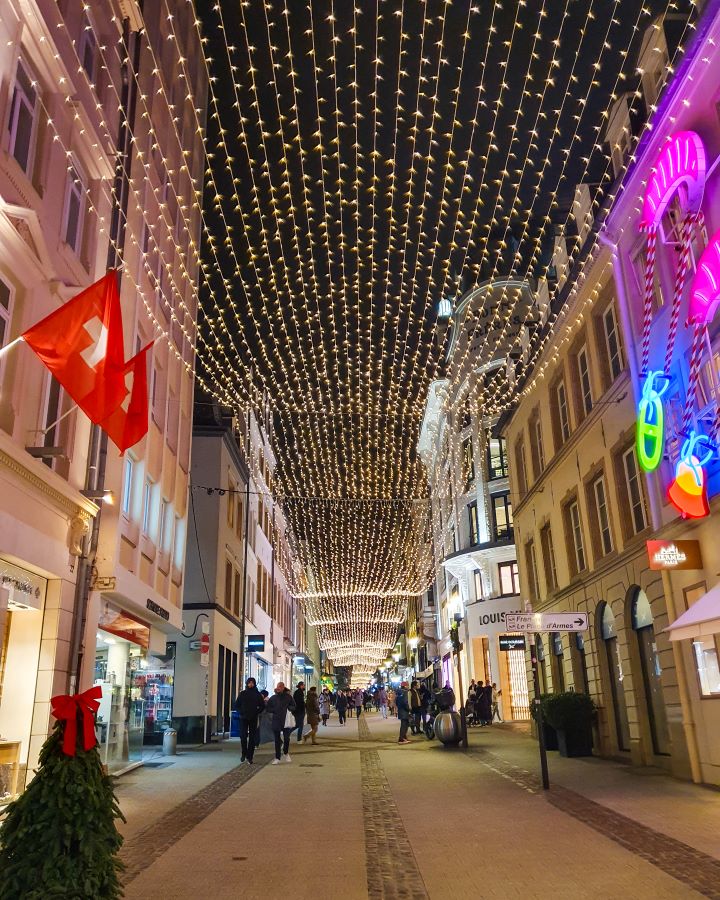 A wide street in Luxembourg City. On either side of the street are buildings and running down the street above are hundreds of fairy lights with people walking underneath. Make the Most of Christmas in Luxembourg City