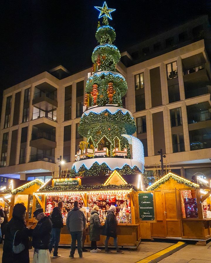 A large Christmas tree in Luxembourg City. The tree is fake and has levels with different decorations on each level. The bottom level even has a train moving around the outside. Under it are some Christmas huts and people walking around. Make the Most of Christmas in Luxembourg City
