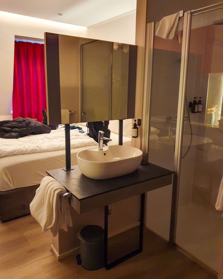 Inside our hotel room in Trier, Germany. There's a bed with some red curtains behind it. In the foreground is a sink with a mirror above it and a shower. See the Amazing Christmas Markets in Trier, Germany