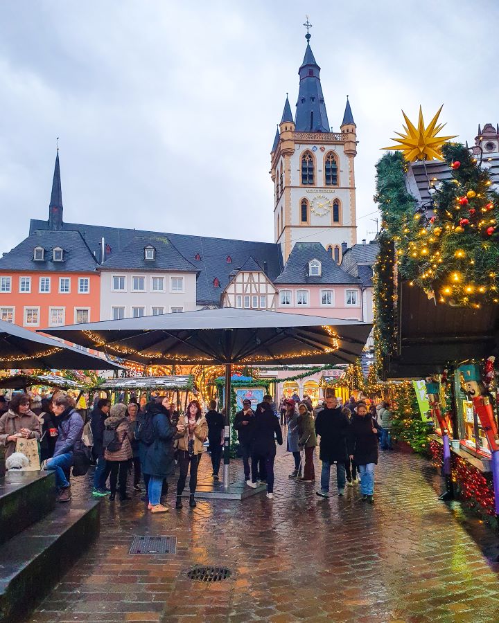 In a Christmas market in Trier, Germany. On the right are wooden huts with garlands and lights on them and on the left are big umbrellas with people walking under them. Behind are some big coloured European buildings in orange, pink and white with a church tower with a black spire coming off it. See the Amazing Christmas Markets in Trier, Germany