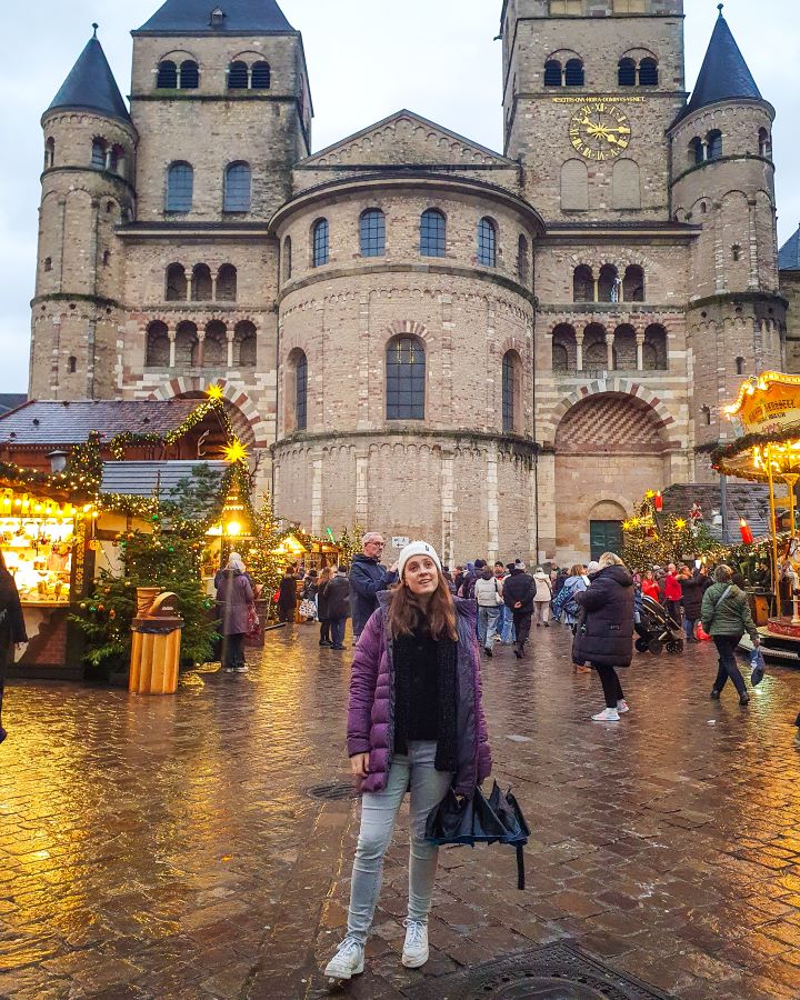 Krissie standing in front of a huge stone cathedral in Trier, Germany. The cathedral has lots of arched windows with 4 towers and a big round bit in the middle. Between Krissie and the cathedral are Christmas huts with lots of lights on them and the side of a carousel on the right with people walking around looking at everything. See the Amazing Christmas Markets in Trier, Germany