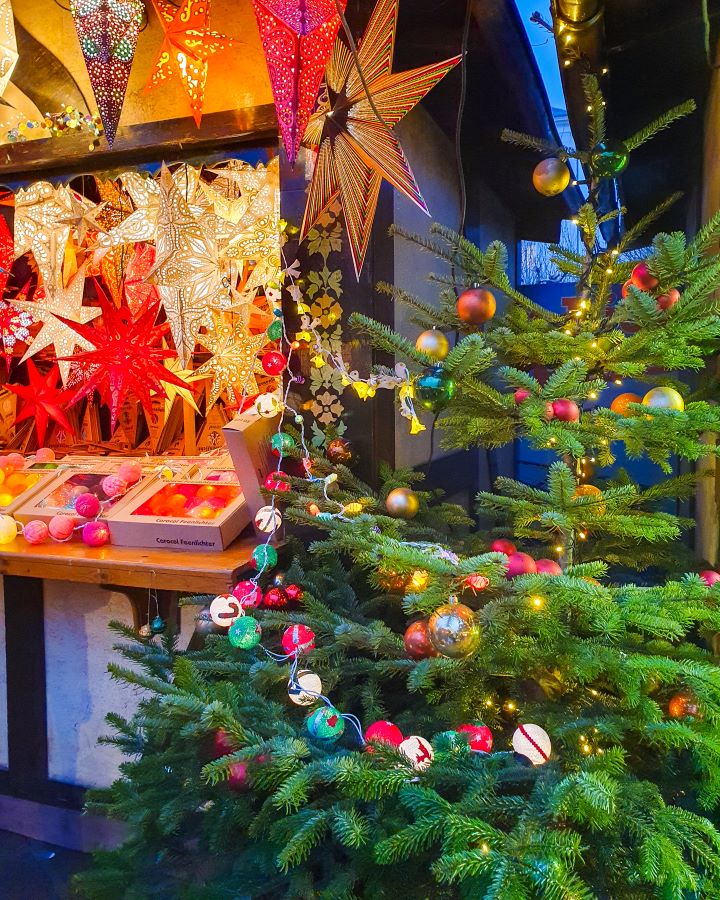 See the Amazing Christmas Markets in Trier, Germany. A real Christmas tree on the right with coloured baubles on it and on the left is a wooden hut selling light up star decorations.