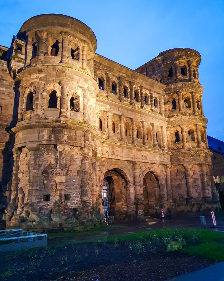 The front of Porta Nigra in Trier, Germany. It's a big stone gate with two rounded sides and a bit in the middle with two archways for people to walk through. See the Amazing Christmas Markets in Trier, Germany