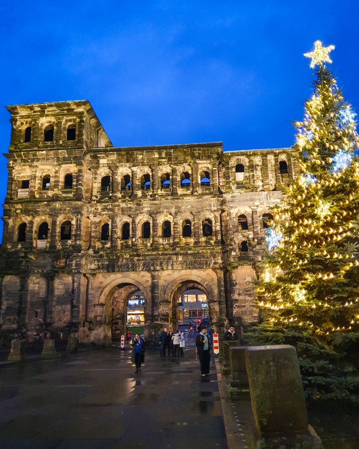 The back of Porta Nigra in Trier, Germany. It's a big stone gate with lots of windows in it and two archways in the bottom for people to walk through. There's people walking under it and a big Christmas tree with a star on top on the right. See the Amazing Christmas Markets in Trier, Germany