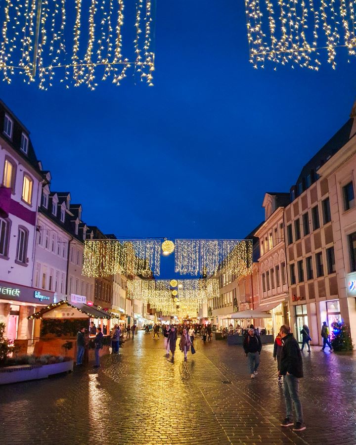 A big main street in Trier, Germany. There's buildings on either side with lights going between them above the walkway. There's people walking around and some Christmas huts too. See the Amazing Christmas Markets in Trier, Germany