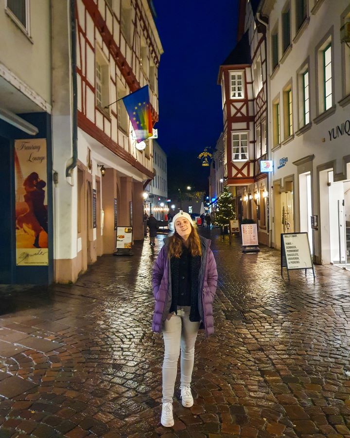Krissie standing on a cobblestone street in Trier, Germany. Behind her are cute German buildings with red beams on the white buildings and some shop boards out of the front of the shops. See the Amazing Christmas Markets in Trier, Germany