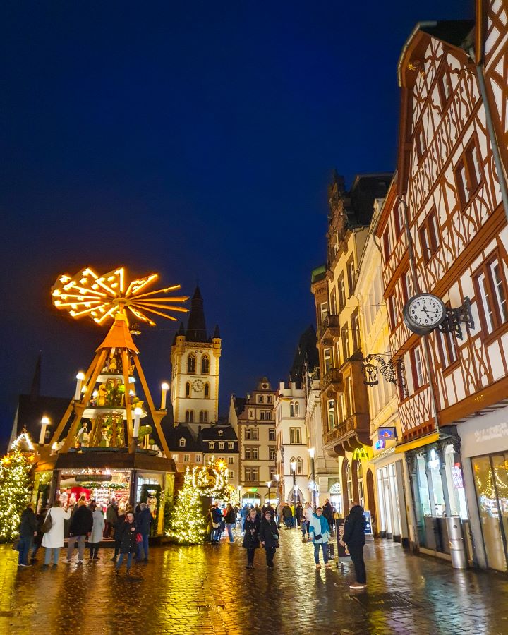 The same Christmas market in Trier, Germany but when its dark outside so everything is lit up. On the right are gorgeous white and brown buildings in a German style. On the left is a structure with lights on it and lit Christmas trees under it. There's also a church tower in the background and people walking around. See the Amazing Christmas Markets in Trier, Germany