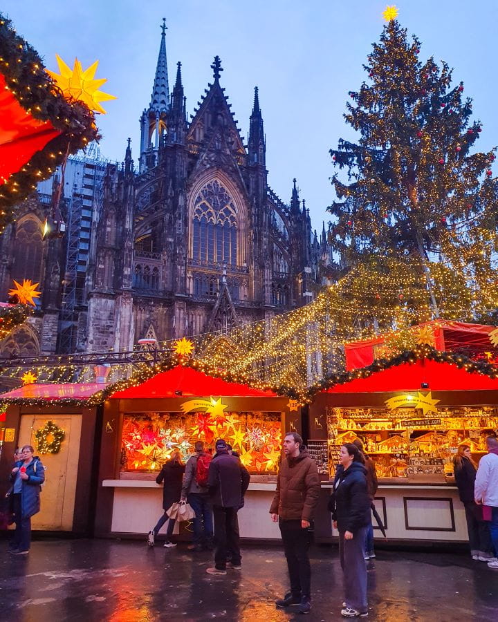 Cathedral Christmas Market Complete Guide to the Magical Cologne Christmas Markets The black gothic looking cathedral behind little huts with red roofs at the Cathedral Christmas Market in Cologne, Germany. The huts are selling lights and other crafts and there's people walking around looking at them. Behind the huts is also a big Christmas tree with a yellow star on top and fairy lights coming off it. Complete Guide to the Magical Cologne Christmas Markets