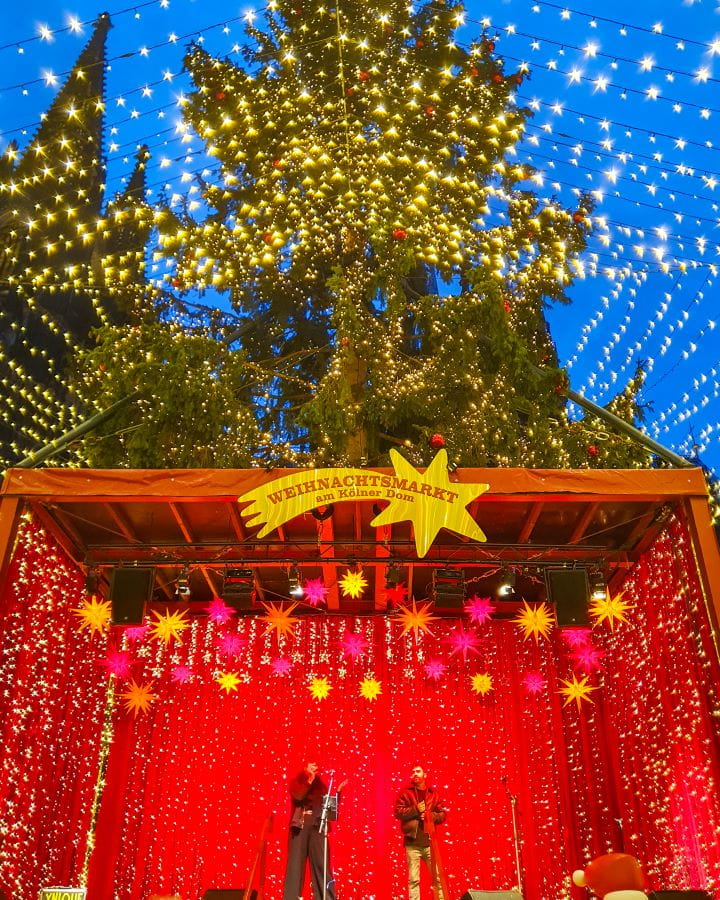 Cathedral Christmas Market Complete Guide to the Magical Cologne Christmas Markets A red stage with fairy lights on it and two people onstage performing at the Cathedral Christmas Market in Cologne, Germany. The stage has a star sign on it that says "Weihnachtsmart am Kolner Dom". Behind that is the big Christmas tree with fairy lights coming off it and you can see the point of the cathedral behind that too. Complete Guide to the Magical Cologne Christmas Markets