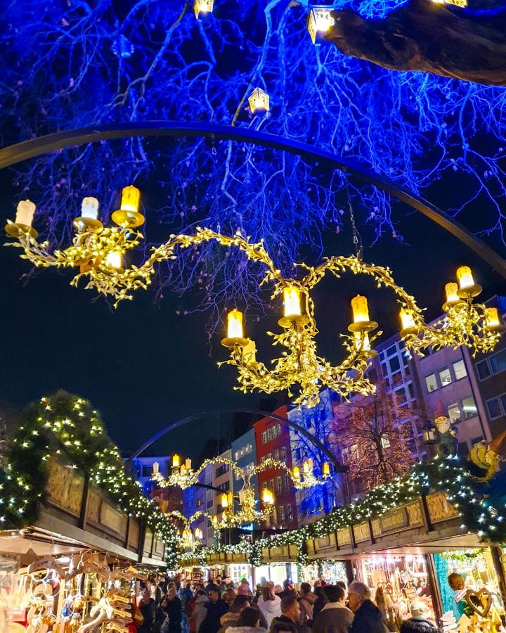 Alter Markt Complete Guide to the Magical Cologne Christmas Markets Looking at a row of Christmas huts with green garlands on top of them with fairy lights in them too at Alter Markt in Cologne Germany. Above the huts are candles and a tree. Behind the huts you can see the coloured buildings of Cologne. Complete Guide to the Magical Cologne Christmas Markets