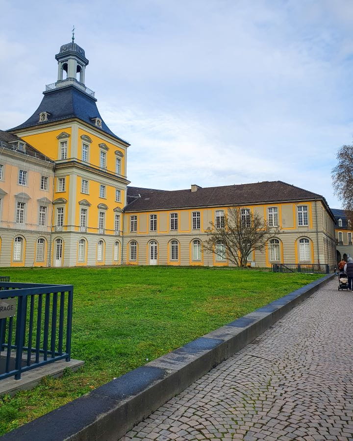 Kurfürstliches Schloss Bonn Germany How To Make The Most of a Day in Bonn from Cologne A yellow and orange building wing of Kurfürstliches Schloss palace which is now the university of Bonn in Germany. In front is some green grass and a cobbled path with a blue sky overhead. How To Make The Most of a Day in Bonn from Cologne