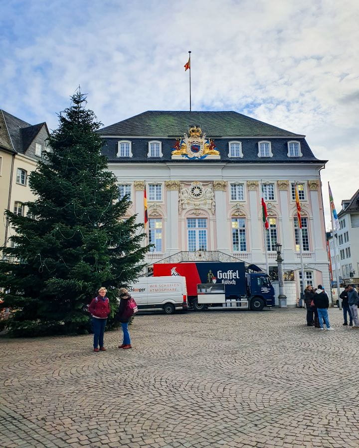 Altes Rathaus Bonn Germany How To Make The Most of a Day in Bonn from Cologne The old town hall in Bonn, Germany. It's a white square building with big windows in front and a black roof with a flag right on top. In front is a staircase and a big Christmas tree with people walking looking at it all. How To Make The Most of a Day in Bonn from Cologne