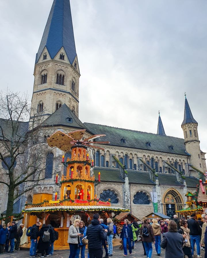 Bonn Minster Münsterplatz Bonn Germany How To Make The Most of a Day in Bonn from Cologne A main square in Bonn, Germany. There's Christmas markets in the square and lots of people walking around. Behind is a big church, Bonn Minster, made of white brick and a dark roof with 3 turrets coming off it. How To Make The Most of a Day in Bonn from Cologne