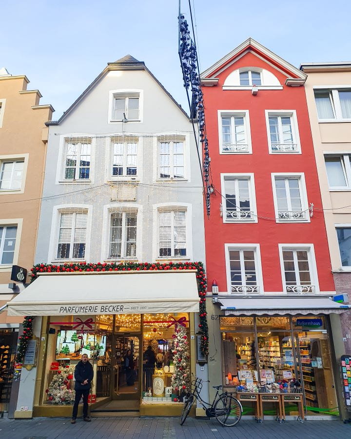 Bonn Germany How To Make The Most of a Day in Bonn from Cologne How To Make The Most of a Day in Bonn from Cologne. Some old buildings in Bonn, Germany. There's an orange one, a light blue one and a red one with shops underneath selling perfume and books. There's also some Christmas decorations around and the sky is a light blue.