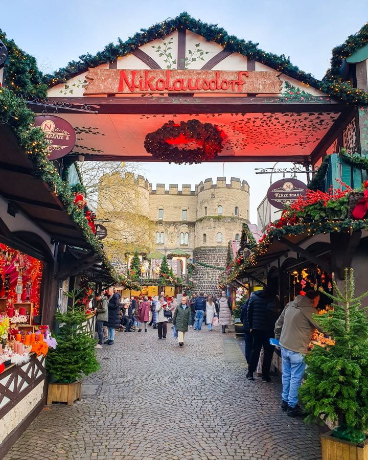 Nikolausdorf Market Complete Guide to the Magical Cologne Christmas Markets The entrance to Nikolausdorf Market in Cologne, Germany. There's a sign over the entrance that says Nikolausolorf. On either side are huts with green garlands on and behind you can see a big stone gate with turrets at the top. There's people walking around and the ground is cobbled. Complete Guide to the Magical Cologne Christmas Markets