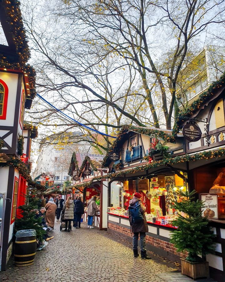 Nikolausdorf Market Complete Guide to the Magical Cologne Christmas Markets Little white and black huts with green garlands running along the top at Nikolausdorf Market in Cologne Germany. There's people walking around as well as little trees, barrels and bigger trees in the background. Complete Guide to the Magical Cologne Christmas Markets