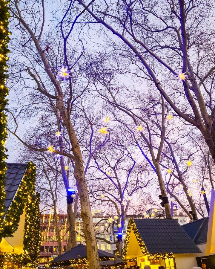 Markt der Engel Complete Guide to the Magical Cologne Christmas Markets The tops of white huts with pointed roofs at a Markt der Engel in Cologne, Germany. The trees above have little star light decorations in them and the sky is purple and blue. Complete Guide to the Magical Cologne Christmas Markets