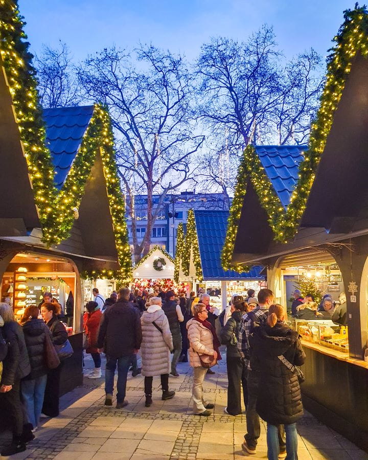 Markt der Engel Complete Guide to the Magical Cologne Christmas Markets Neat black and white huts with pointed roofs and green garlands with fairy lights lining them in Markt der Engel in Cologne, Germany. There's lots of people looking around with trees and the twilight sky in the background. Complete Guide to the Magical Cologne Christmas Markets