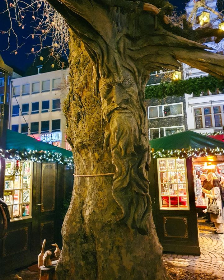Alter Markt Complete Guide to the Magical Cologne Christmas Markets A tree with a wooden detail on it of an old mans face which looks very mythical and cool at Alter Markt in Cologne, Germany. Behind the tree are some huts with green roofs and fairy lights. Complete Guide to the Magical Cologne Christmas Markets