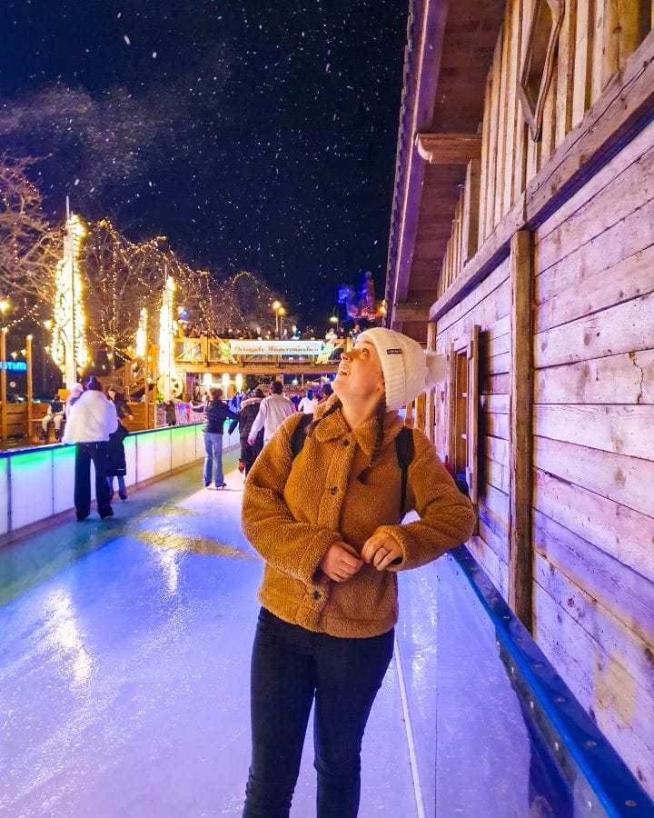 Heumarkt Complete Guide to the Magical Cologne Christmas Markets Krissie standing on the ice rink and looking up at the fake snow over her at Heumarkt in Cologne, Germany. It's night and you can see the snow against the sky and behind her are people skating. Complete Guide to the Magical Cologne Christmas Markets