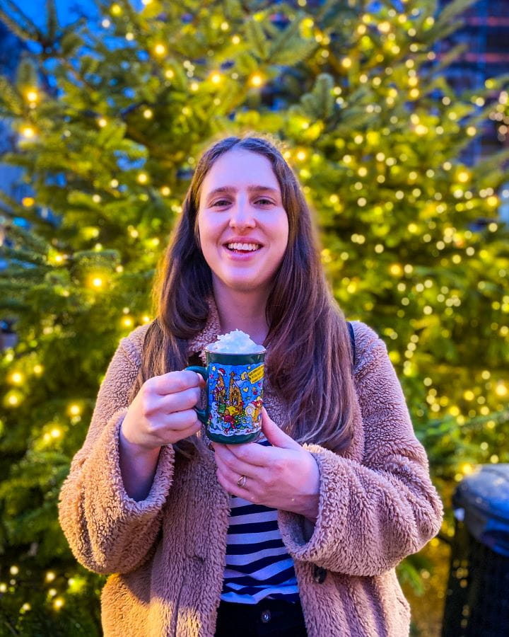 Cathedral Christmas Market Complete Guide to the Magical Cologne Christmas Markets Complete Guide to the Magical Cologne Christmas Markets. Katherine smiling at the camera and holding a green mug with a cartoon picture on it and whipped cream on top at the Cathedral Christmas Market in Cologne, Germany. Behind her the background is blurred but it's a Christmas tree with lights on it.