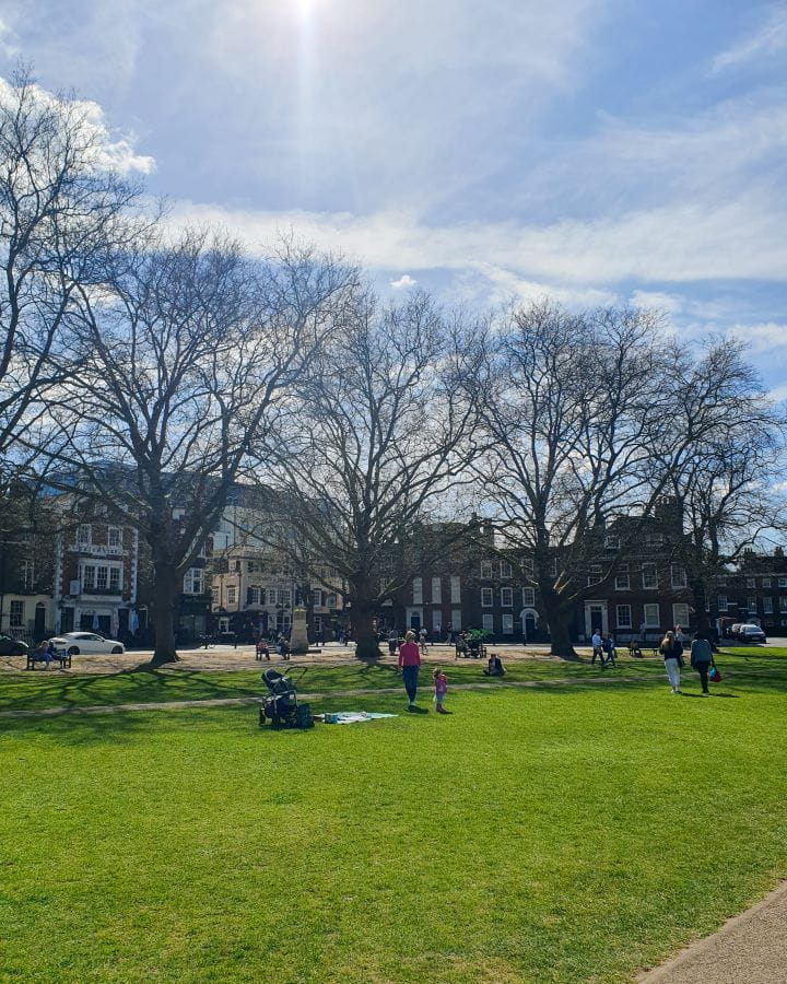 Richmond Green London Less Touristy Places in London for Return Visitors A green patch of grass in Richmond, London. There's bare trees on the outskirts, people on the grass and small buildings behind it all. Less Touristy Places in London for Return Visitors