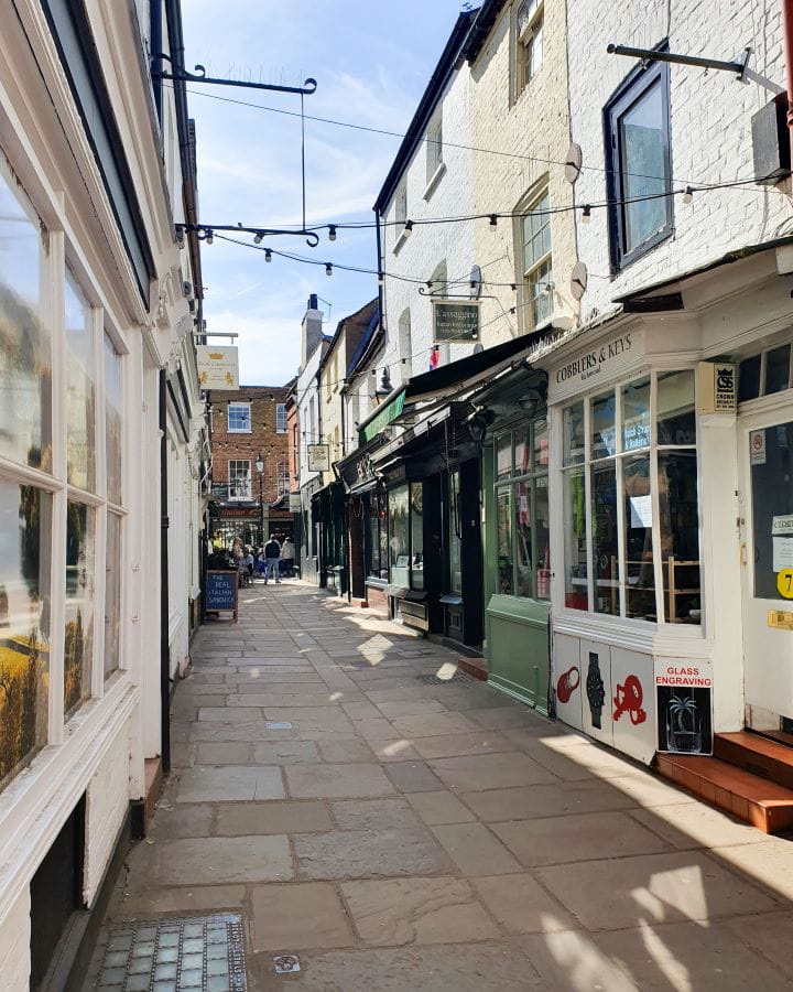 Richmond Ted Lasso London Less Touristy Places in London for Return Visitors A laneway in Richmond, London. The ground is paved and the shops on either side are cute, with apartments on top of them and fairy lights above the path. Less Touristy Places in London for Return Visitors