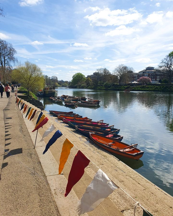 Richmond Thames River London Less Touristy Places in London for Return Visitors Along the river Thames in Richmond, London. There's bunting flags lining the water, little red boats on the edge, and a path with people walking down it on the left. There's also lots of trees along the water and the sky above is blue. Less Touristy Places in London for Return Visitors