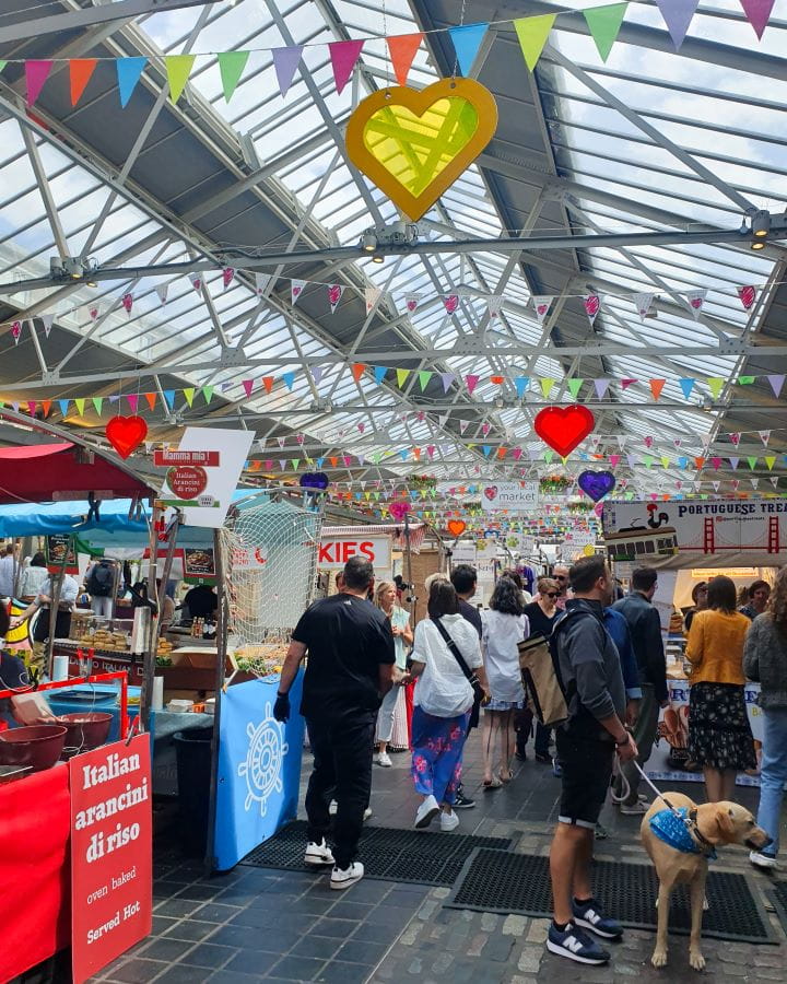 Greenwich Market London Less Touristy Places in London for Return Visitors Under the glass panelled roof of Greenwich Market in London. There's coloured bunting and decorations hanging from the roof and lots of stalls under it with people walking around. Less Touristy Places in London for Return Visitors
