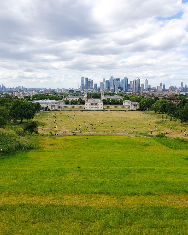 Greenwich Park London Less Touristy Places in London for Return Visitors The view from Greenwich Park back to London. There's a big green hill with lots of people picnicking at the bottom. There's green trees on either side of the hill and a big white palace at the bottom with a view of London behind it all. Less Touristy Places in London for Return Visitors