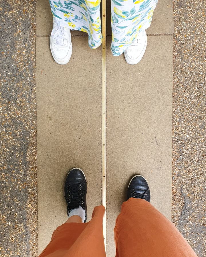 Greenwich Park London Less Touristy Places in London for Return Visitors The photo is taken of our feet from above. Katherine's orange pants and black shoes with Krissie's white, yellow and green pants and white shoes standing on a path with a metal line running between their feet in Greenwich, London. Less Touristy Places in London for Return Visitors