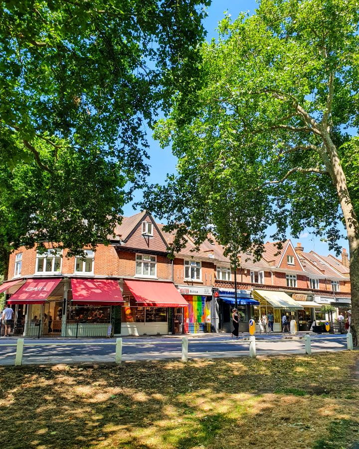 Barnes London Less Touristy Places in London for Return Visitors A cute set of shops in Barnes, London. There's coloured awnings over shops on the bottom and above are orange brick housing. There's a road between the shops and a grassy patch and lots of green trees overhead. Less Touristy Places in London for Return Visitors